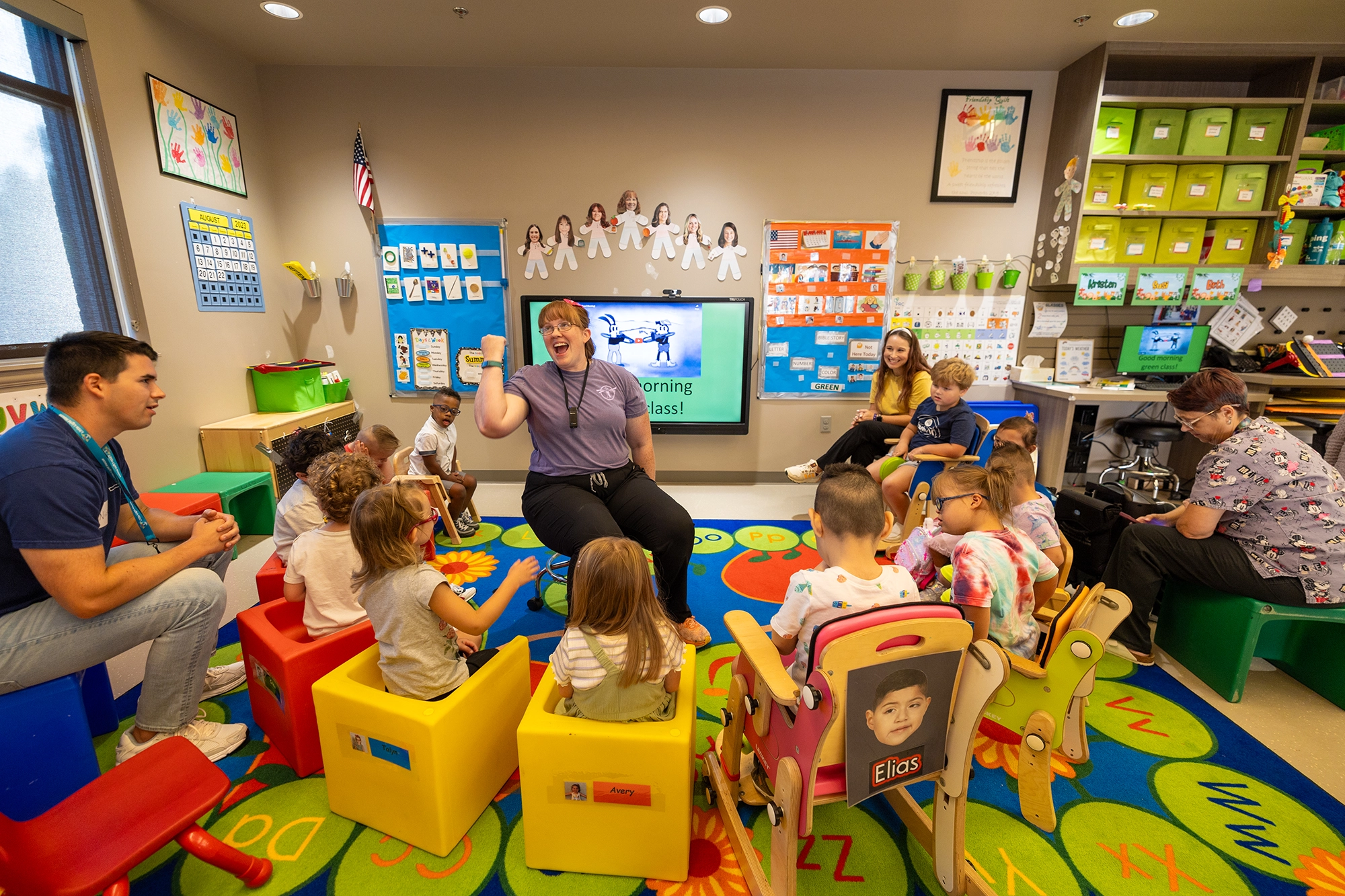 Wide shot of Little Light House classroom with children with disabilities sitting in adapted chairs and teacher is in the center excited to start the day